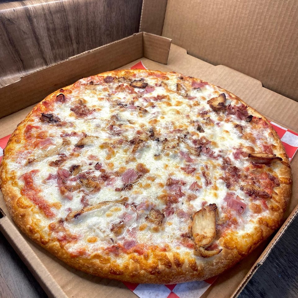 A freshly baked pizza with melted cheese, ham, and chicken in a cardboard delivery box.
