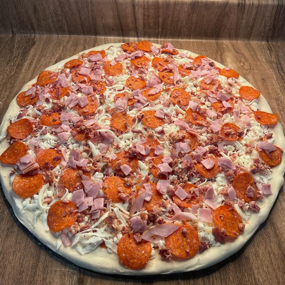 Unbaked pizza with pepperoni, ham, and cheese on a wooden counter.