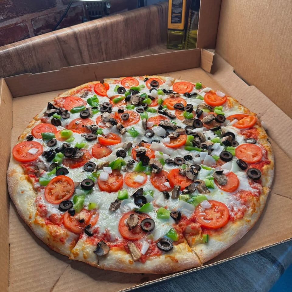A whole pizza in a cardboard box, topped with tomatoes, green peppers, onions, mushrooms, and olives.