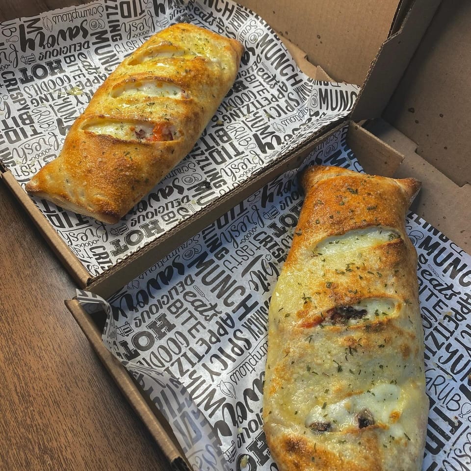 Two golden-brown calzones with melted cheese and herbs, in cardboard boxes with patterned paper.