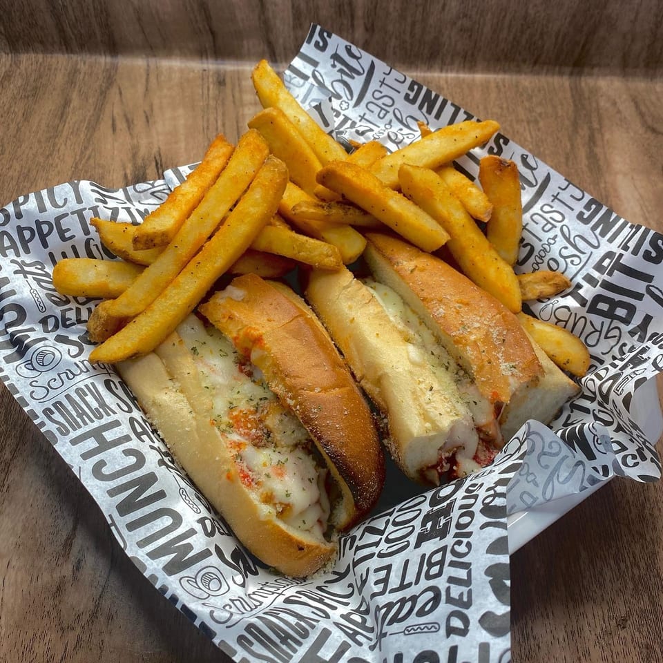 Two cheesy subs with marinara and fries in a basket lined with branded paper.