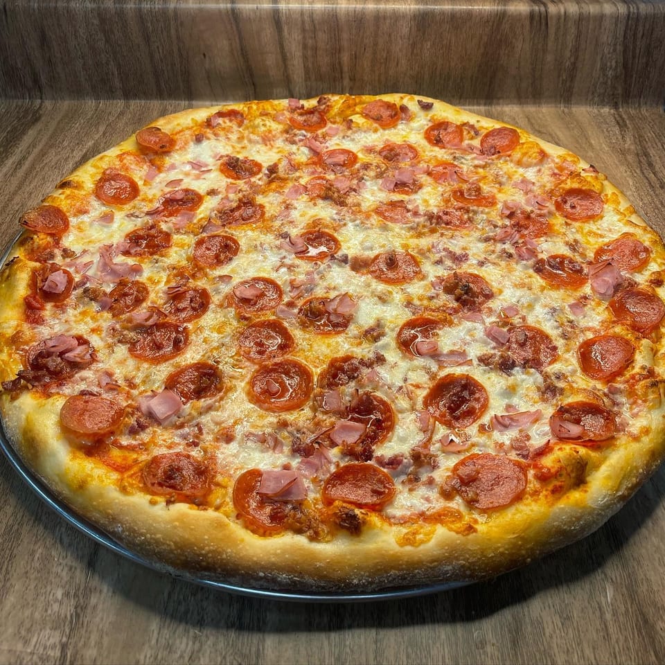 A large, freshly baked pizza with pepperoni, ham, and sausage on a wooden surface.